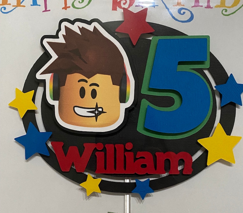 Pode incluir: Um topo de bolo preto e branco com um personagem Roblox, um n&uacute;mero 5 azul, estrelas amarelas e o nome William em vermelho.