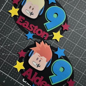 Pode incluir: Dois toppers para bolo de anivers&aacute;rio em preto e branco com um personagem de desenho animado usando fones de ouvido e o n&uacute;mero 9. Os toppers t&ecirc;m os nomes Easton e Alden escritos em vermelho e bord&ocirc;.