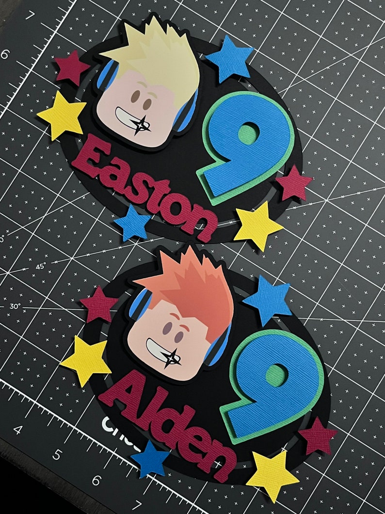 Pode incluir: Dois toppers para bolo de anivers&aacute;rio em preto e branco com um personagem de desenho animado usando fones de ouvido e o n&uacute;mero 9. Os toppers t&ecirc;m os nomes Easton e Alden escritos em vermelho e bord&ocirc;.