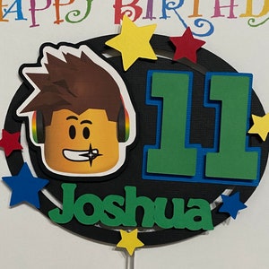 Pode incluir: Um topo de bolo preto e branco com um personagem de desenho animado usando fones de ouvido, o n&uacute;mero 11 e o nome Joshua. O topo &eacute; decorado com estrelas coloridas.