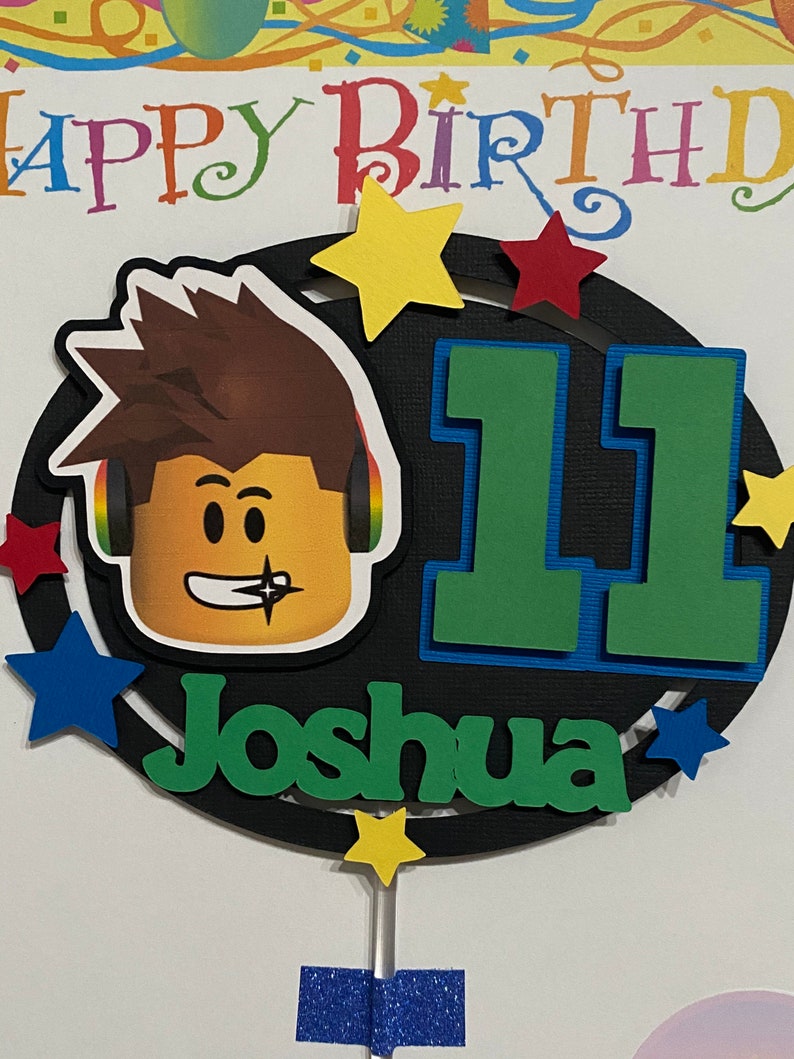 Pode incluir: Um topo de bolo preto e branco com um personagem de desenho animado usando fones de ouvido, o n&uacute;mero 11 e o nome Joshua. O topo &eacute; decorado com estrelas coloridas.