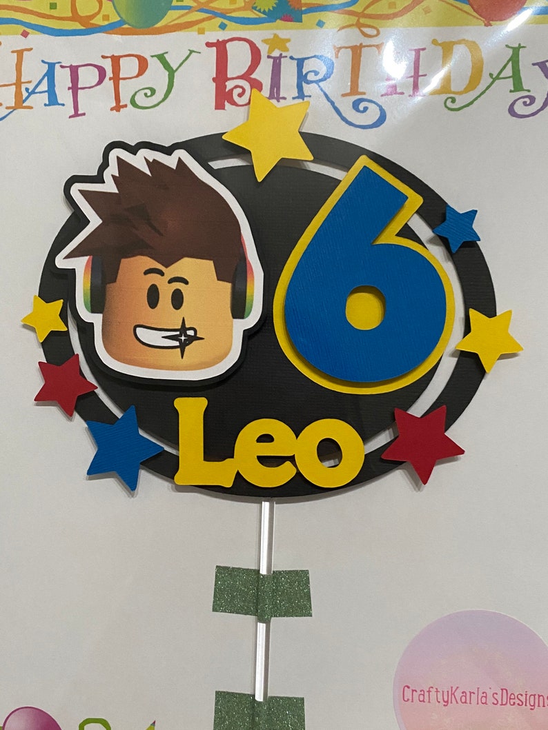 Pode incluir: Um topo de bolo com um personagem Roblox com um n&uacute;mero 6 azul e amarelo, o nome Leo e estrelas amarelas em um fundo preto.
