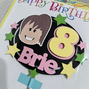 Pode incluir: Um cart&atilde;o de anivers&aacute;rio com um c&iacute;rculo preto com um n&uacute;mero 8 amarelo, um nome rosa "Brie" e uma menina de desenho animado com cabelo castanho. O cart&atilde;o tem uma borda colorida com o texto "Feliz anivers&aacute;rio" em rosa, amarelo, azul e verde.