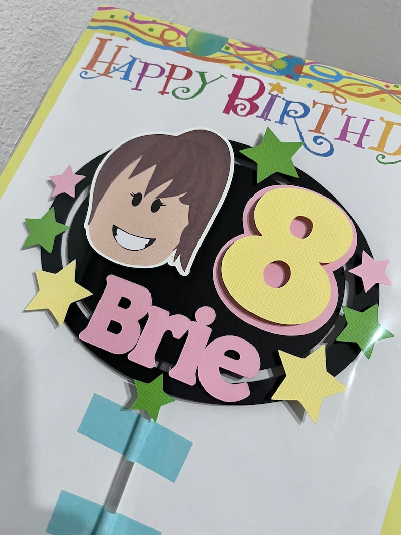 Pode incluir: Um cart&atilde;o de anivers&aacute;rio com um c&iacute;rculo preto com um n&uacute;mero 8 amarelo, um nome rosa "Brie" e uma menina de desenho animado com cabelo castanho. O cart&atilde;o tem uma borda colorida com o texto "Feliz anivers&aacute;rio" em rosa, amarelo, azul e verde.