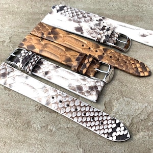 Puede incluir: Varias correas de reloj con un patrón de piel de serpiente en tonos blancos, marrones y negros. Cada correa tiene una hebilla plateada. Las correas están hechas de un material texturizado y están dispuestas en diagonal.