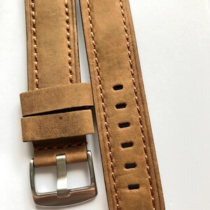 May include: A pair of light brown leather watch straps with a silver-colored buckle. The straps have a stitched border and several holes for adjustment. The leather has a textured appearance.