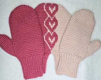 Könnte beinhalten: Drei gestrickte Fäustlinge in Rosa- und Beigetönen. Der Fäustling links ist einfarbig tiefrosa. Der mittlere Fäustling hat ein rosa-weißes Herzmuster. Der Fäustling rechts ist hellbeige. Alle Fäustlinge haben ein strukturiertes Strickmuster.