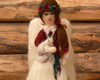 May include: A felt angel ornament with a white dress, red jacket, and a green and red floral crown. The angel is holding a white dove in its hands.