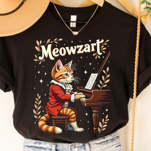 K&ouml;nnte beinhalten: Schwarzes T-Shirt mit einem Aufdruck eines Katers, der Klavier spielt. Der Text "Meowzart" befindet sich &uuml;ber dem Kater.