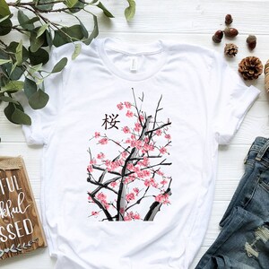 Puede incluir: Camiseta blanca con un gr&aacute;fico de un &aacute;rbol de cerezo en flor negro y rosa y la palabra japonesa "桜" (Sakura) impresa en la parte delantera. La camiseta est&aacute; colocada sobre una superficie blanca con un par de pantalones vaqueros azules y un letrero de madera que dice "Grateful Thankful Blessed".