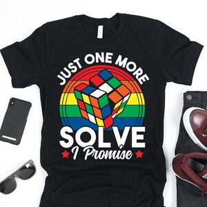 Pode incluir: Camiseta preta com um círculo arco-íris em torno de um cubo de Rubik. O texto "Just One More Solve I Promise" está escrito em branco na camiseta.