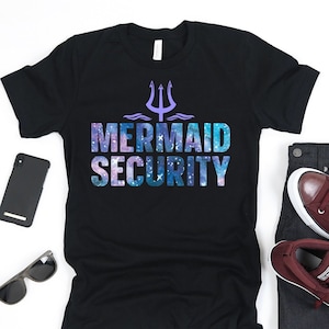 Puede incluir: Camiseta negra con estampado de galaxia y el texto "Mermaid Security" en morado y azul. Un tridente se encuentra sobre el texto.