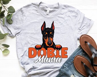 May include: A grey t-shirt with a Doberman Pinscher peeking over the top. The text "Doberman Mama" is printed in orange below the dog.