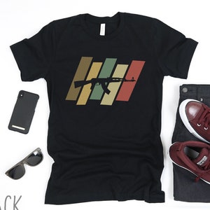 May include: Black t-shirt featuring a retro-style graphic of a rifle with a multi-coloured striped background.