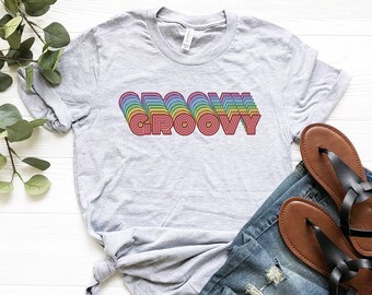 Puede incluir: Una camiseta gris con la palabra "GROOVY" impresa en colores arcoíris. La camiseta está colocada sobre una superficie blanca con un par de sandalias marrones y unos vaqueros azules.