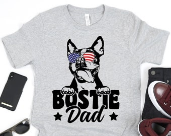 May include: A gray t-shirt with a black and white illustration of a Boston Terrier wearing sunglasses with an American flag design. The text "Bostie Dad" is printed below the dog with two stars on either side.