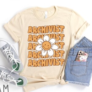May include: A cream colored t-shirt with the word "Archivist" repeated five times in a retro font. Each word is surrounded by small flower graphics. A smiling daisy with a yellow face is centered on the shirt.