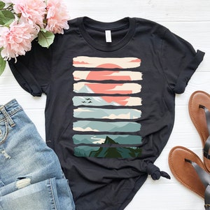 Puede incluir: Camiseta negra con un estampado gr&aacute;fico de un paisaje de monta&ntilde;a con un sol rojo y p&aacute;jaros volando por encima. El gr&aacute;fico est&aacute; formado por pinceladas horizontales.