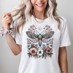 May include: White t-shirt with a graphic of a moth surrounded by pink and white flowers. The moth has intricate details and is surrounded by a floral design.