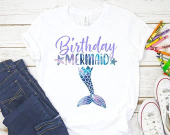 May include: White t-shirt with the text "Birthday Mermaid" in purple lettering with a sparkly purple and blue mermaid tail graphic. The shirt is laid out on a white wooden surface with a pair of white sneakers, blue jeans, and a box of crayons.