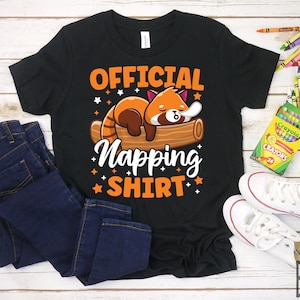 Może przedstawiać: Czarny T-shirt z czerwonym pandą śpiącym na kłodzie. Na koszulce widnieje napis "Official Napping Shirt" z gwiazdami wokół tekstu.