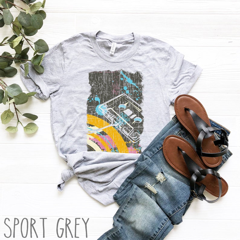 May include: A grey t-shirt featuring a vintage-style graphic of a cassette tape with colourful accents. The shirt is laid flat on a white wooden surface with a pair of brown sandals and blue jeans.