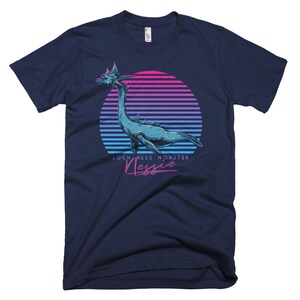 Puede incluir: Una camiseta azul marino con un gr&aacute;fico retro del monstruo del Lago Ness, Nessie, frente a una puesta de sol rosa y azul. El texto "Loch Ness Monster Nessie" est&aacute; impreso debajo del gr&aacute;fico.