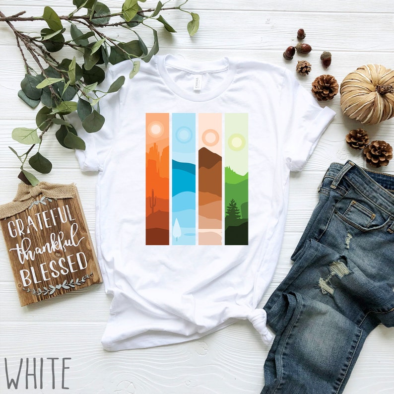 May include: White t-shirt with a graphic of four vertical stripes, each depicting a different landscape scene: a desert, a lake, a mountain range, and a forest. The text "Grateful Thankful Blessed" is visible on a wooden sign in the background.