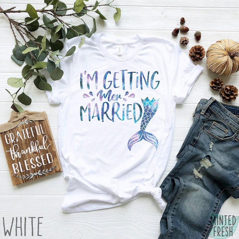 Peut inclure: T-shirt blanc avec le texte "I'm getting mer-married" en lettres scintillantes bleues et violettes. Une queue de sir&egrave;ne avec un imprim&eacute; galaxie est &agrave; droite du texte.