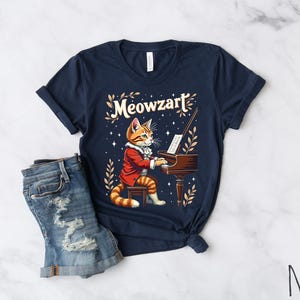 K&ouml;nnte beinhalten: Ein marineblaues T-Shirt mit einem Aufdruck eines Katers, der Klavier spielt. Der Text "Meowzart" ist auf dem Shirt gedruckt.