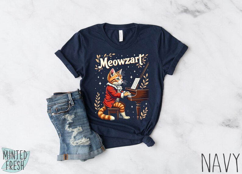 K&ouml;nnte beinhalten: Ein marineblaues T-Shirt mit einem Aufdruck eines Katers, der Klavier spielt. Der Text "Meowzart" ist auf dem Shirt gedruckt.