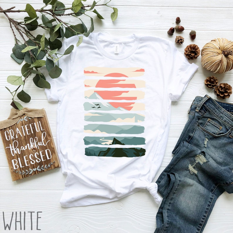 Puede incluir: Camiseta blanca con un dise&ntilde;o gr&aacute;fico de una cordillera con una puesta de sol en el fondo. El dise&ntilde;o est&aacute; formado por rayas horizontales de diferentes colores, incluyendo verde, azul y naranja. El texto "GRATEFUL thankful BLESSED" es visible en un letrero de madera en la imagen.