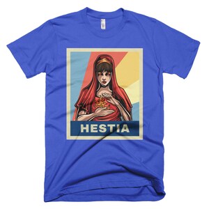K&ouml;nnte beinhalten: Ein blaues T-Shirt mit einem Retro-Poster-Design, das eine Frau mit roten Haaren und einem roten Gewand zeigt, die eine Flamme in ihren H&auml;nden h&auml;lt. Der Text des Posters lautet "Hestia".