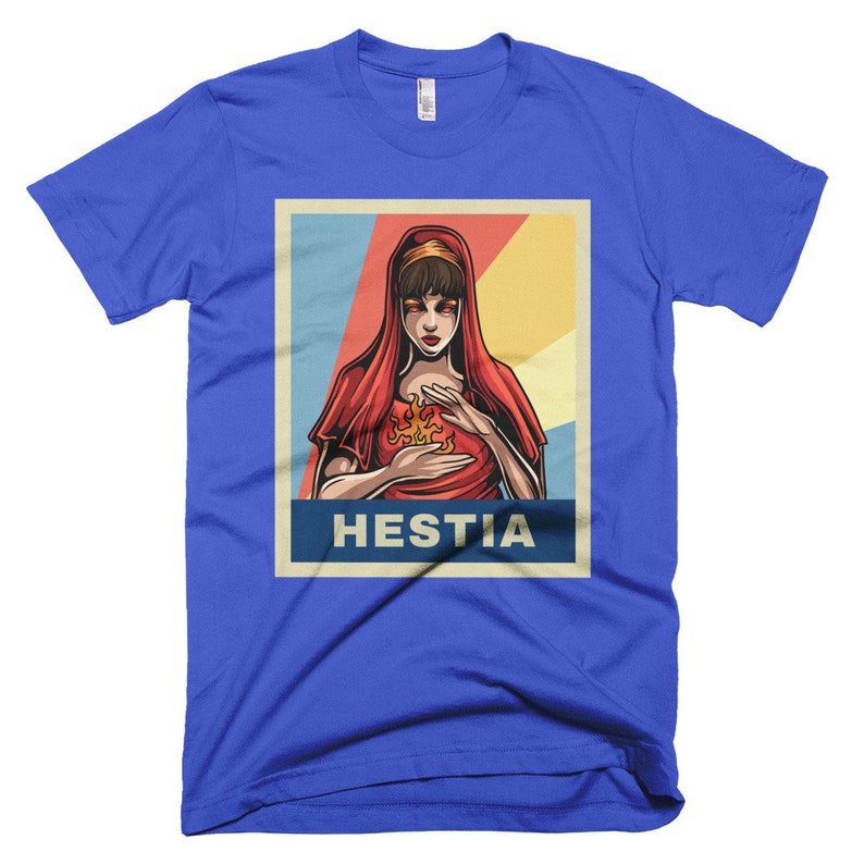 K&ouml;nnte beinhalten: Ein blaues T-Shirt mit einem Retro-Poster-Design, das eine Frau mit roten Haaren und einem roten Gewand zeigt, die eine Flamme in ihren H&auml;nden h&auml;lt. Der Text des Posters lautet "Hestia".