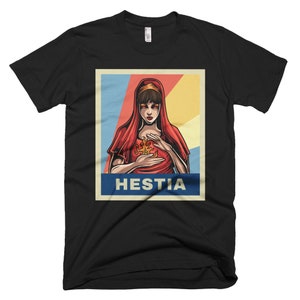 K&ouml;nnte beinhalten: Ein schwarzes T-Shirt mit einer Retro-Grafik einer Frau mit roten Haaren und einem roten Kleid, die eine Flamme in ihren H&auml;nden h&auml;lt. Die Grafik ist vor einem blauen, wei&szlig;en und gelben Hintergrund platziert. Der Text "Hestia" ist in Wei&szlig; unter der Grafik gedruckt.