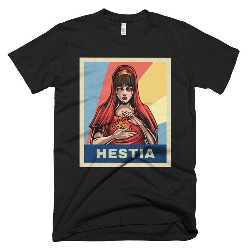 K&ouml;nnte beinhalten: Ein schwarzes T-Shirt mit einer Retro-Grafik einer Frau mit roten Haaren und einem roten Kleid, die eine Flamme in ihren H&auml;nden h&auml;lt. Die Grafik ist vor einem blauen, wei&szlig;en und gelben Hintergrund platziert. Der Text "Hestia" ist in Wei&szlig; unter der Grafik gedruckt.