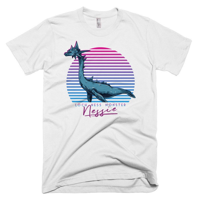 Puede incluir: Camiseta blanca con un gr&aacute;fico de estilo retro del Monstruo del Lago Ness, Nessie, en colores azul y verde, con una puesta de sol rosa y morada detr&aacute;s. El texto "Loch Ness Monster Nessie" est&aacute; impreso debajo del gr&aacute;fico.