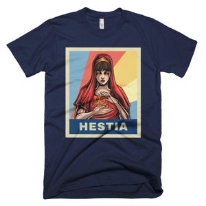 K&ouml;nnte beinhalten: Ein marineblaues T-Shirt mit einem Vintage-Poster-Design, das eine Frau mit roten Haaren und einem roten Gewand zeigt, die eine Flamme in ihren H&auml;nden h&auml;lt. Der Text des Posters lautet "Hestia".