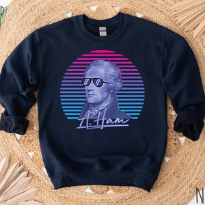 May include: A navy blue sweatshirt featuring a retro-style graphic of Alexander Hamilton wearing sunglasses against a pink, blue and purple sunset. The text "A. Ham" is written beneath the graphic.