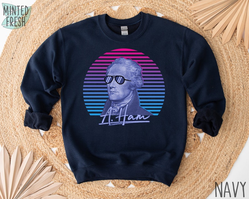 May include: A navy blue sweatshirt featuring a retro-style graphic of Alexander Hamilton wearing sunglasses against a pink, blue and purple sunset. The text "A. Ham" is written beneath the graphic.