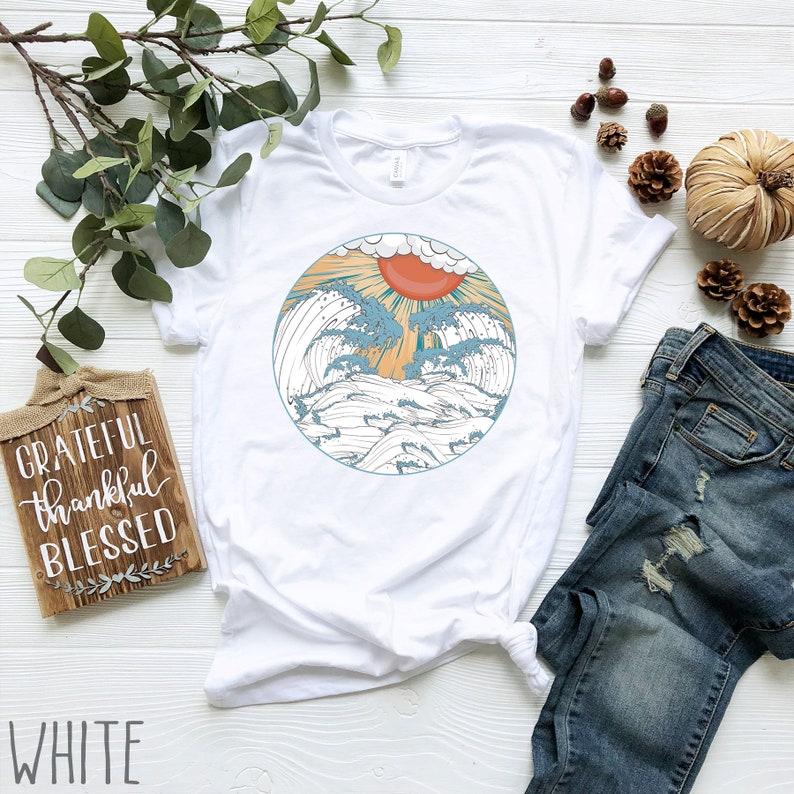 May include: White t-shirt featuring a graphic of a sun setting over waves within a circle. The graphic is in shades of blue, orange, and yellow.