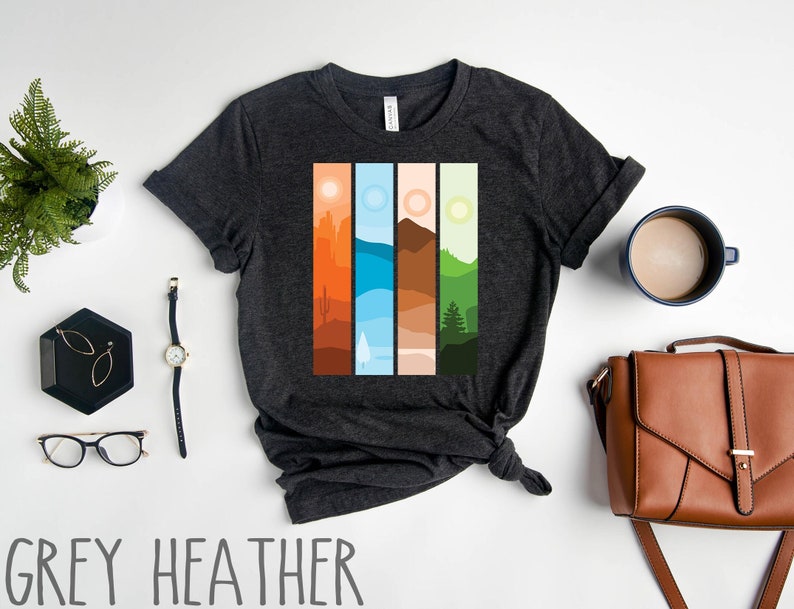 May include: A dark heather grey t-shirt with a graphic design of four vertical panels depicting different landscapes. The panels are in shades of orange, blue, brown, and green.