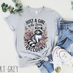 May include: A gray t-shirt with the text "Just a girl who loves raccoons" and a cartoon raccoon surrounded by flowers.
