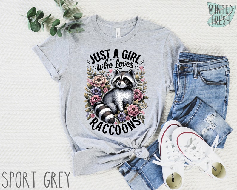 May include: A gray t-shirt with the text "Just a girl who loves raccoons" and a cartoon raccoon surrounded by flowers.