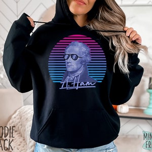 May include: Black hoodie featuring a retro-style graphic of Alexander Hamilton wearing sunglasses against a pink and blue sunset backdrop. The text "A. Ham" is written in a stylized font below the graphic.