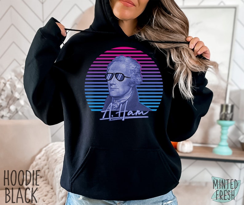 May include: Black hoodie featuring a retro-style graphic of Alexander Hamilton wearing sunglasses against a pink and blue sunset backdrop. The text "A. Ham" is written in a stylized font below the graphic.
