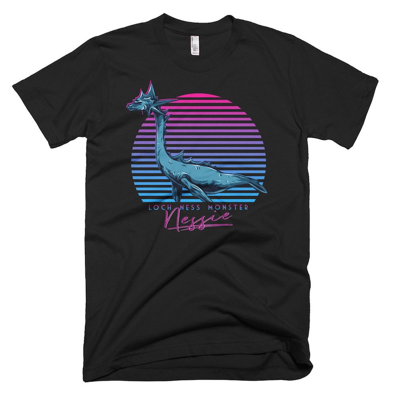 Puede incluir: Una camiseta negra con un gr&aacute;fico de estilo retro del monstruo del Lago Ness, Nessie, nadando en una puesta de sol rosa y azul. El texto "Loch Ness Monster Nessie" est&aacute; impreso debajo del gr&aacute;fico.