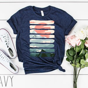 Puede incluir: Una camiseta azul marino con un estampado gr&aacute;fico de un paisaje de monta&ntilde;a con una puesta de sol. El gr&aacute;fico est&aacute; formado por rayas horizontales en varios tonos de verde, azul y naranja.