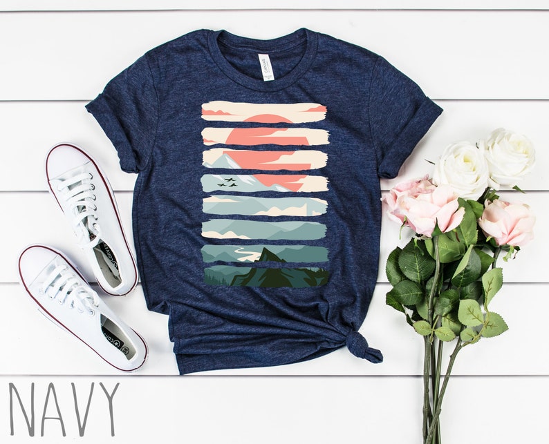 Puede incluir: Una camiseta azul marino con un estampado gr&aacute;fico de un paisaje de monta&ntilde;a con una puesta de sol. El gr&aacute;fico est&aacute; formado por rayas horizontales en varios tonos de verde, azul y naranja.