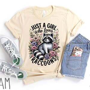May include: A cream colored t-shirt with the text "Just a girl who loves raccoons" in black lettering. The text is surrounded by a floral design with a raccoon in the center.